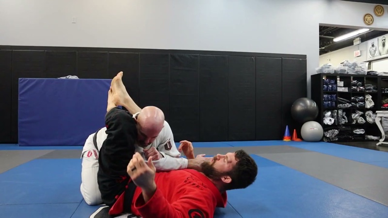 The attacker in a white gi finishes an armbar from closed guard, hips off the mat, while the partner in a red gi lies on their back.