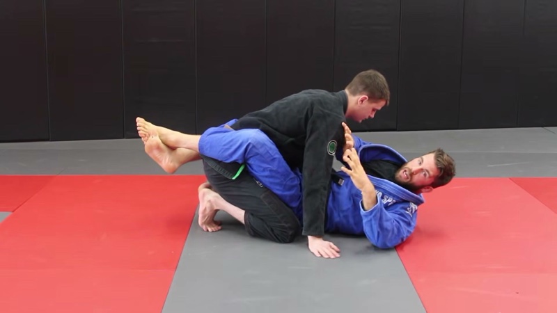 The bottom player in a blue gi locks closed guard around a kneeling partner in a black gi and talks through a detail.