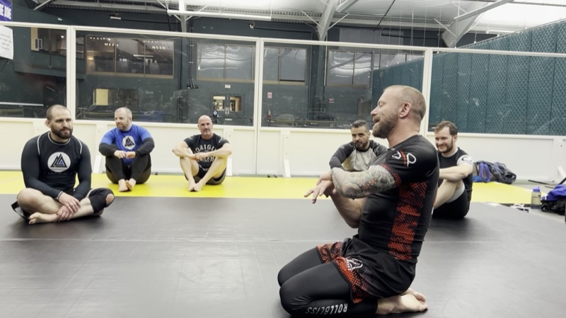 An instructor sits on the mats explaining a point to a circle of seated students.